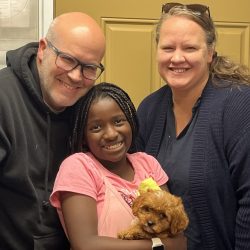 Three adults and a young girl smile at the camera while the girl holds a small brown puppy.