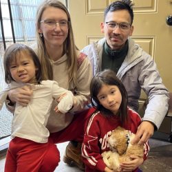 Four people pose indoors near a door; a child in red holds two small puppies on her lap.