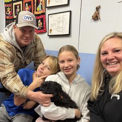 Four people sit indoors, smiling at the camera, with a black dog in the center held by the girl in the white hoodie.