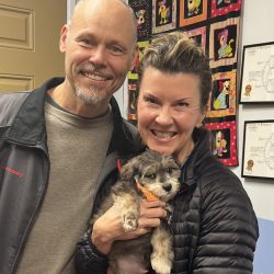 Two adults smile while holding a small, fluffy puppy inside a room with colorful art and certificates on the wall.