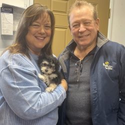 Two people smile at the camera; one holds a small black and white puppy in front of a tan door indoors.