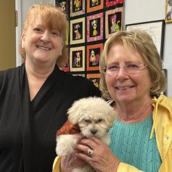 Two women smiling indoors, one holding a small white puppy wearing a red bandana, with a colorful quilt in the background.
