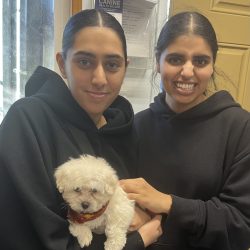 Two women in black hoodies stand indoors, one holding a small white dog wearing a red scarf.