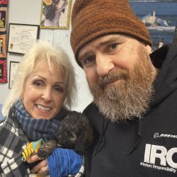 A smiling woman and a bearded man hold a small black puppy indoors, surrounded by colorful wall art.