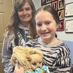 A woman and a girl smile at the camera; the girl is holding a small dog wrapped in blankets.