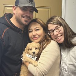 Three people smile at the camera as the woman in the middle holds a small tan puppy in front of a tan door.