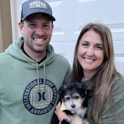 Two smiling adults stand together outside, with one holding a small black and white puppy in their arms.