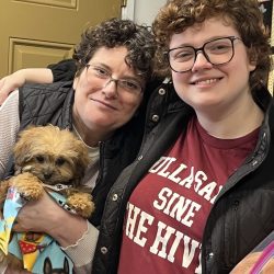Two people smile at the camera; one holds a small dog wrapped in a colorful blanket inside a home.
