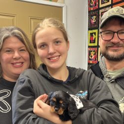 Three adults and a girl pose indoors; the girl holds a small black and brown puppy wrapped in a white blanket.