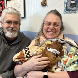 A man and woman sit indoors, smiling, as the woman holds a small puppy wrapped in colorful blankets.