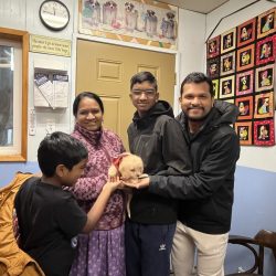 A family of four stands indoors, holding a small puppy and smiling at the camera in a decorated room.
