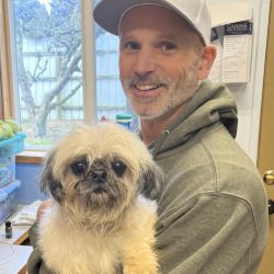 A man in a gray cap and hoodie holds a small, fluffy dog inside a room with a window and papers on the wall.