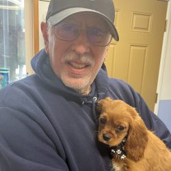 Older man in a navy hoodie and cap holds a small brown puppy with a black collar, standing indoors near a tan door.