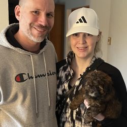 Two adults stand indoors; one holds a small brown puppy. Both are smiling at the camera.