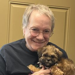 Older woman with short gray hair and glasses holding a small brown puppy, standing in front of a tan door.