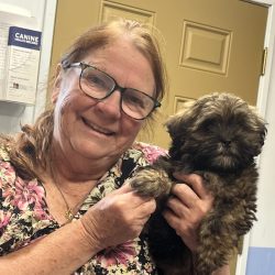 A woman with glasses smiles while holding a small, furry brown puppy in front of a tan door.
